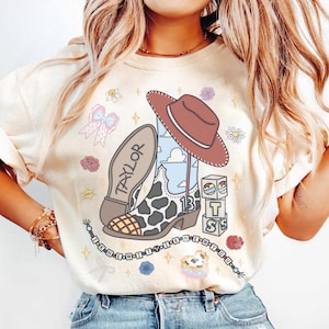 Puede incluir: Una camiseta color crema con un estampado gr&aacute;fico de una bota vaquera con patr&oacute;n de manchas de vaca, un sombrero marr&oacute;n y el nombre Taylor. El dise&ntilde;o incluye bloques con las letras T, S, el n&uacute;mero 13 y un collar de cuentas con el texto I KNEW IT I KNEW YOU.