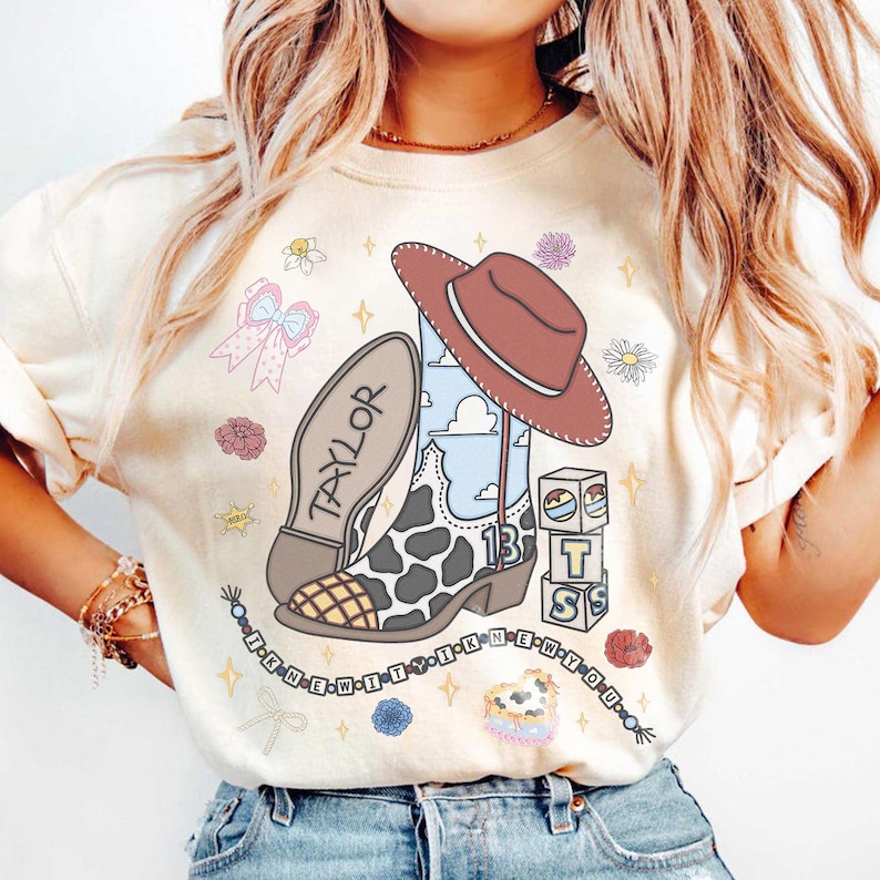 Puede incluir: Una camiseta color crema con un estampado gr&aacute;fico de una bota vaquera con patr&oacute;n de manchas de vaca, un sombrero marr&oacute;n y el nombre Taylor. El dise&ntilde;o incluye bloques con las letras T, S, el n&uacute;mero 13 y un collar de cuentas con el texto I KNEW IT I KNEW YOU.