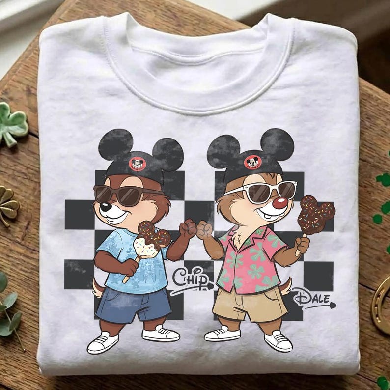 Pu&ograve; includere: Felpa bianca girocollo con un'illustrazione di Chip e Dale con occhiali da sole e cappelli di Topolino. Tengono in mano gelati al cioccolato. Lo sfondo ha un motivo a scacchiera in bianco e nero. Le parole "Chip" e "Dale" sono stampate.