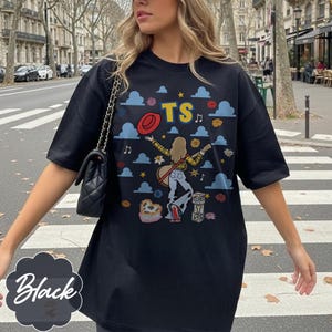 May include: A black short-sleeve t-shirt featuring a graphic print of a person with long hair playing a guitar, surrounded by blue clouds, red hats, flowers, musical notes, and the letters TS. The design includes stacked blocks with TS.