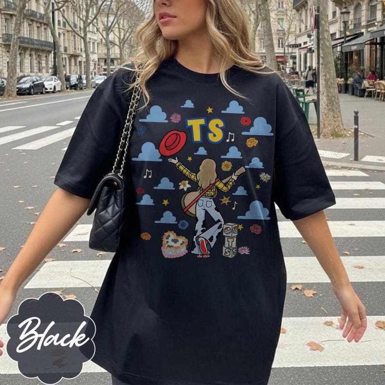 May include: A black short-sleeve t-shirt featuring a graphic print of a person with long hair playing a guitar, surrounded by blue clouds, red hats, flowers, musical notes, and the letters TS. The design includes stacked blocks with TS.