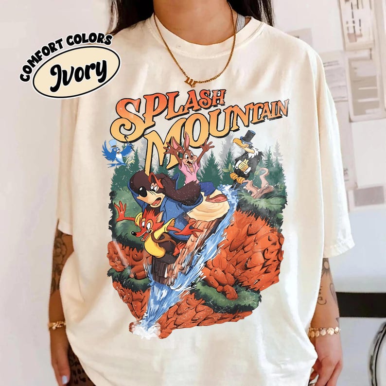 May include: Ivory t-shirt featuring a colourful graphic of a cartoon scene from Splash Mountain. The graphic shows animated characters on a log flume ride, with the words "Splash Mountain" in orange and yellow.