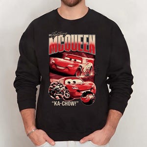 Puede incluir: Sudadera negra con un dise&ntilde;o de Rayo McQueen de la pel&iacute;cula Cars. El dise&ntilde;o incluye el texto "Lightning McQueen" en rojo y blanco, im&aacute;genes del coche de carreras rojo y la frase "KA-CHOW!"