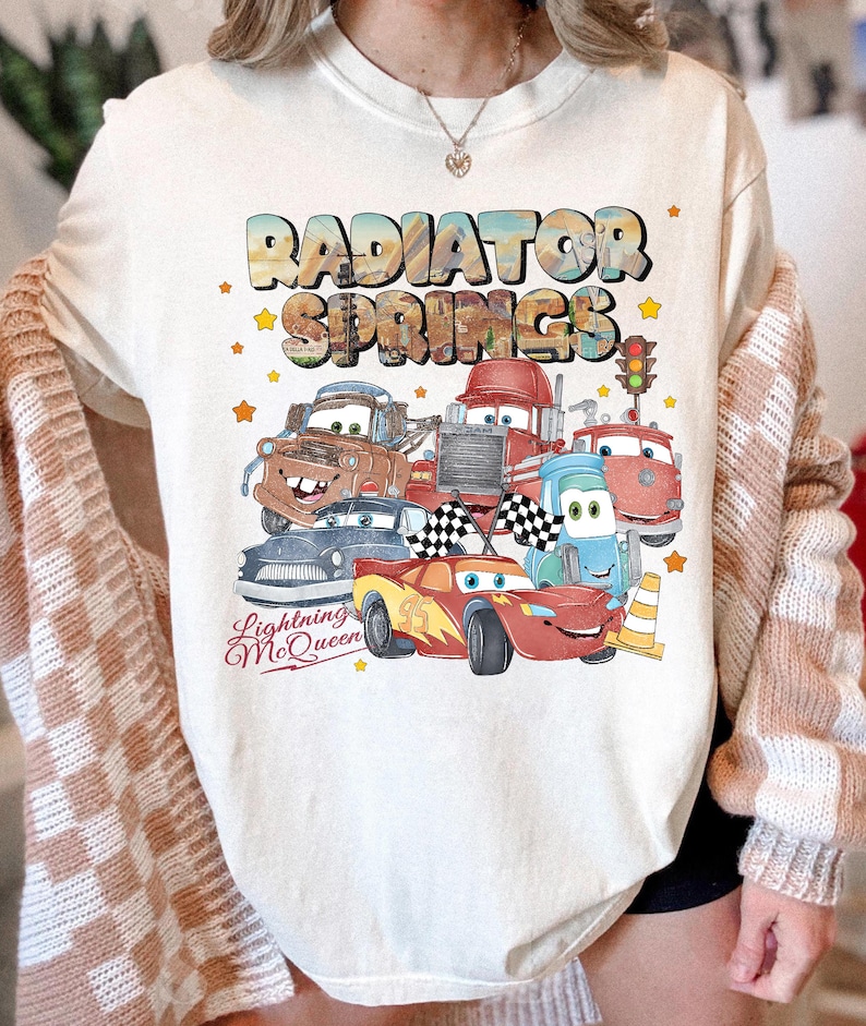 May include: Cream-colored sweatshirt featuring a colorful graphic of characters from the movie Cars, including Lightning McQueen. The text "RADIATOR SPRINGS" and "Lightning McQueen" are also displayed. The design includes stars and a traffic light.