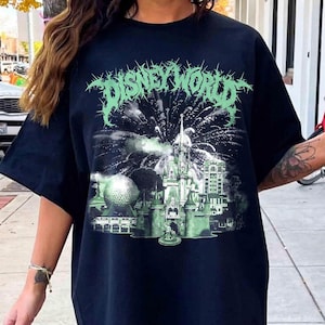 K&ouml;nnte beinhalten: Schwarzes T-Shirt mit einer gr&uuml;n-wei&szlig;en Grafik, die ein Feuerwerk &uuml;ber einem Schloss zeigt, mit dem Text "DISNEY WORLD" in einer stilisierten Schriftart. Das Design umfasst eine golfballf&ouml;rmige Struktur und andere Geb&auml;ude.
