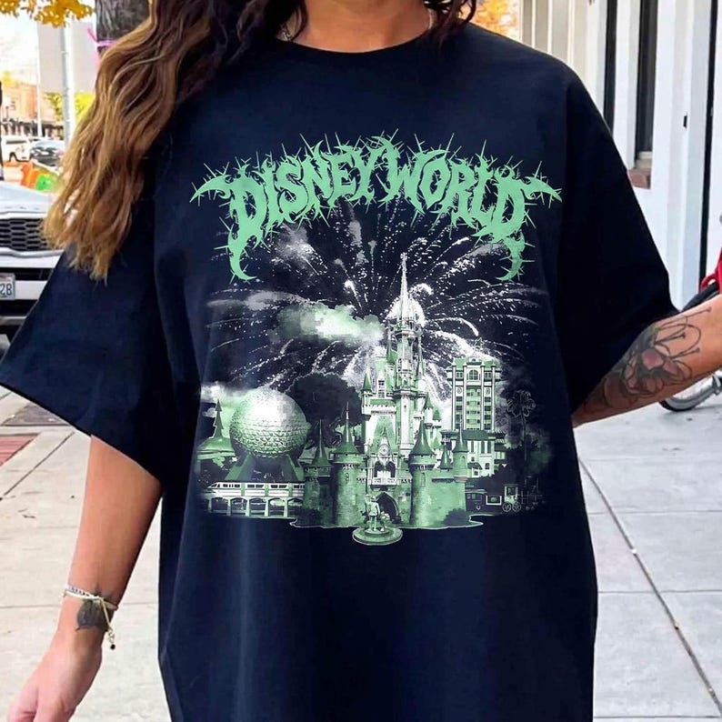 K&ouml;nnte beinhalten: Schwarzes T-Shirt mit einer gr&uuml;n-wei&szlig;en Grafik, die ein Feuerwerk &uuml;ber einem Schloss zeigt, mit dem Text "DISNEY WORLD" in einer stilisierten Schriftart. Das Design umfasst eine golfballf&ouml;rmige Struktur und andere Geb&auml;ude.
