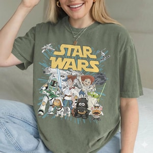 Puede incluir: Camiseta gr&aacute;fica verde oliva de Star Wars con personajes de las franquicias Star Wars y Toy Story. El dise&ntilde;o incluye el logotipo de Star Wars e im&aacute;genes de Darth Vader, Woody y otros personajes.