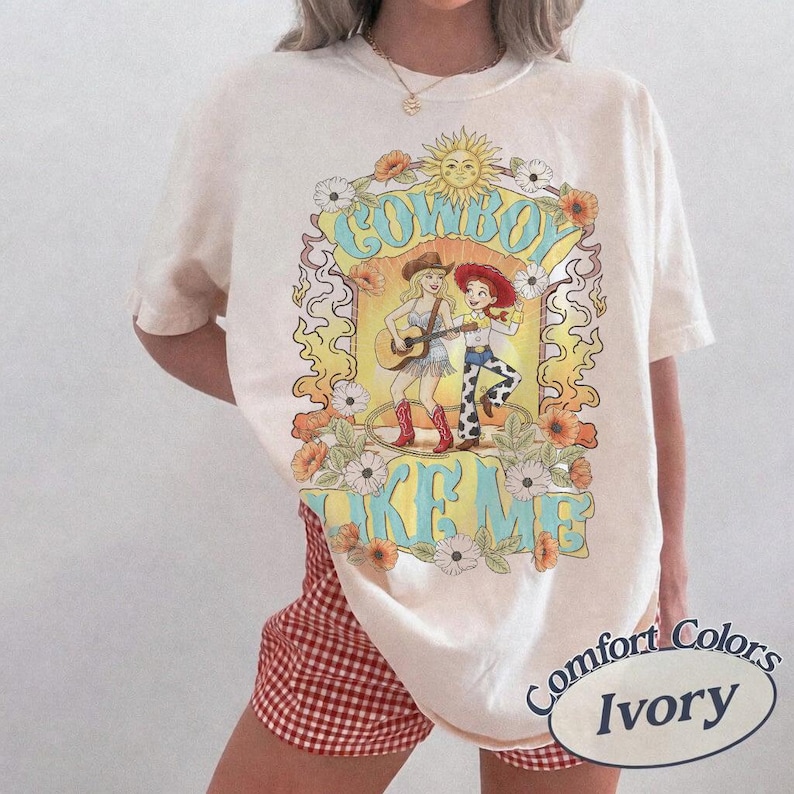 May include: An ivory short-sleeve t-shirt featuring a graphic print of two cartoon characters in western attire. The design includes the text COWBOY LIKE ME, a sun, and floral accents in shades of yellow, orange, blue, and brown. The shirt is worn with red and white checkered shorts.