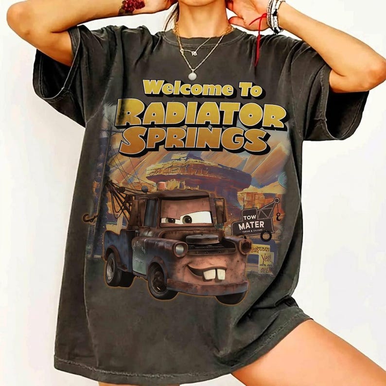 Pu&ograve; includere: T-shirt grigio scuro con la scritta "Welcome To Radiator Springs" e un'illustrazione di Cricchetto del film Cars. La grafica include un paesaggio desertico e un cartello con la scritta "Tow Mater".