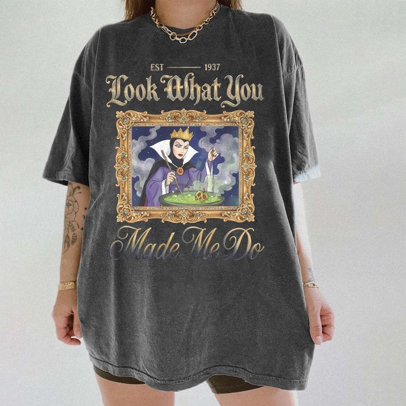 May include: Dark gray t-shirt with a framed image of a cartoon villain stirring a potion. The text on the shirt reads "EST 1937 Look What You Made Me Do". The shirt is oversized and has short sleeves.