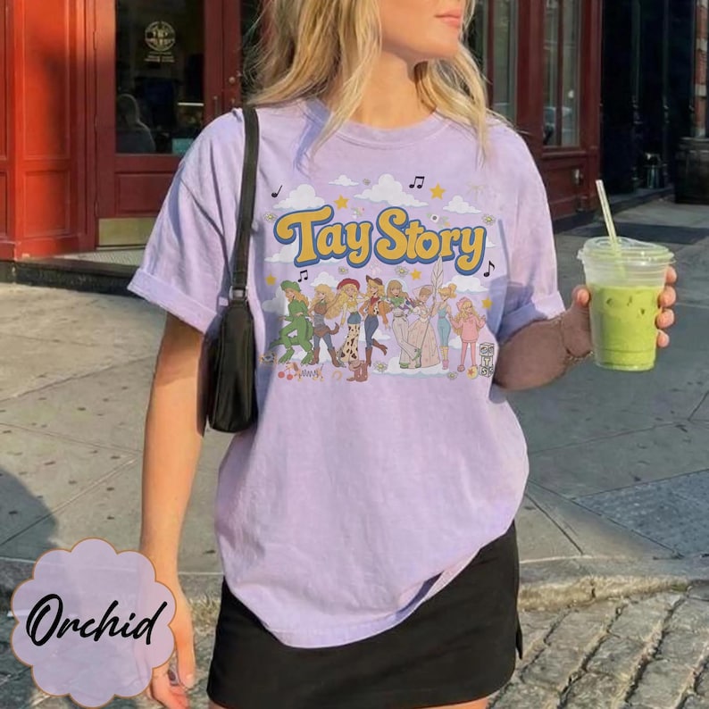May include: A light purple short-sleeve t-shirt featuring the text Tay Story in yellow with a blue outline. Below the text is a graphic illustration of several figures in various outfits, surrounded by clouds, stars, and musical notes.