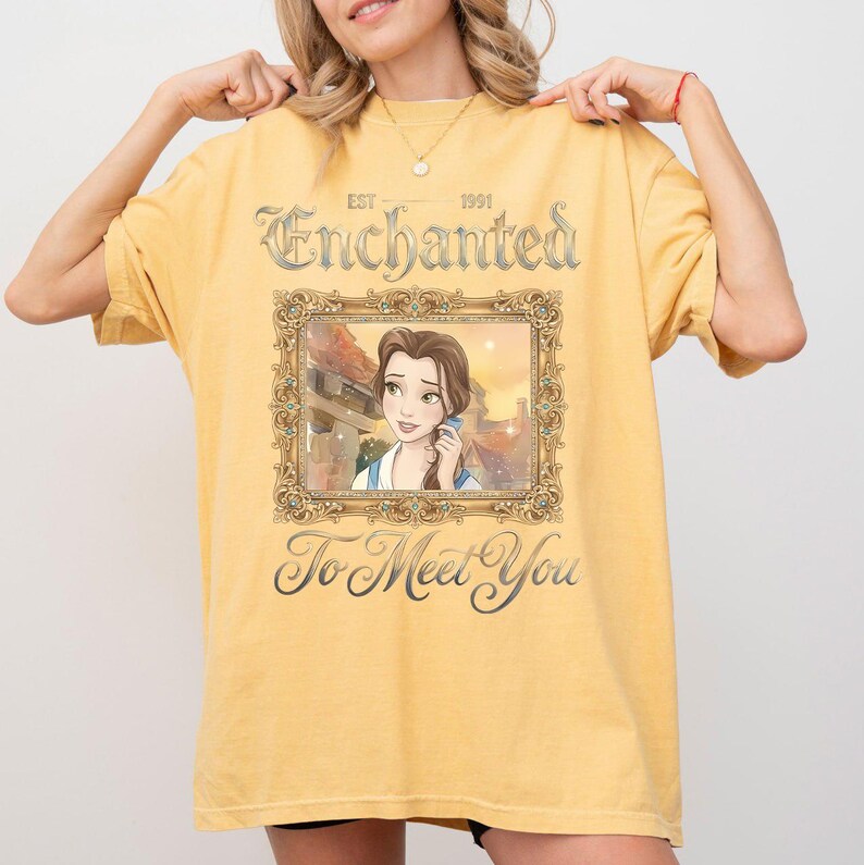 May include: Mustard yellow t-shirt with the text "Enchanted To Meet You" and a framed image of a cartoon character. The shirt has a vintage aesthetic, with the year 1991 printed above the framed image.