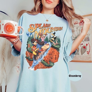 May include: Pale blue t-shirt featuring the words "Splash Mountain" in orange and yellow. The graphic shows cartoon characters on a water ride. A white and orange mug with a sun design is held in the left hand.