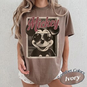 May include: A brown t-shirt featuring a vintage-style graphic of Mickey Mouse wearing sunglasses. The word "Mickey" is printed above the character. The shirt is labeled "Comfort Colors Ivory" at the bottom.