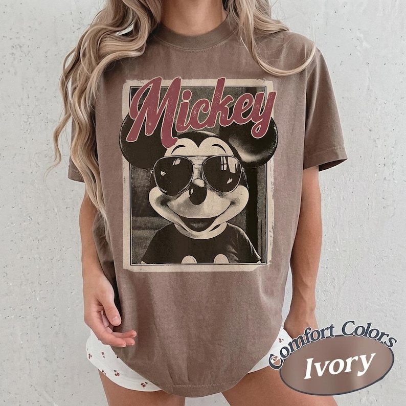 May include: A brown t-shirt featuring a vintage-style graphic of Mickey Mouse wearing sunglasses. The word "Mickey" is printed above the character. The shirt is labeled "Comfort Colors Ivory" at the bottom.