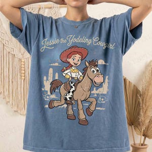 May include: A blue short-sleeve t-shirt featuring a graphic print of Jessie the Yodeling Cowgirl riding Bullseye the horse. The design includes a desert landscape with cacti and the text Jessie the Yodeling Cowgirl in a script font.