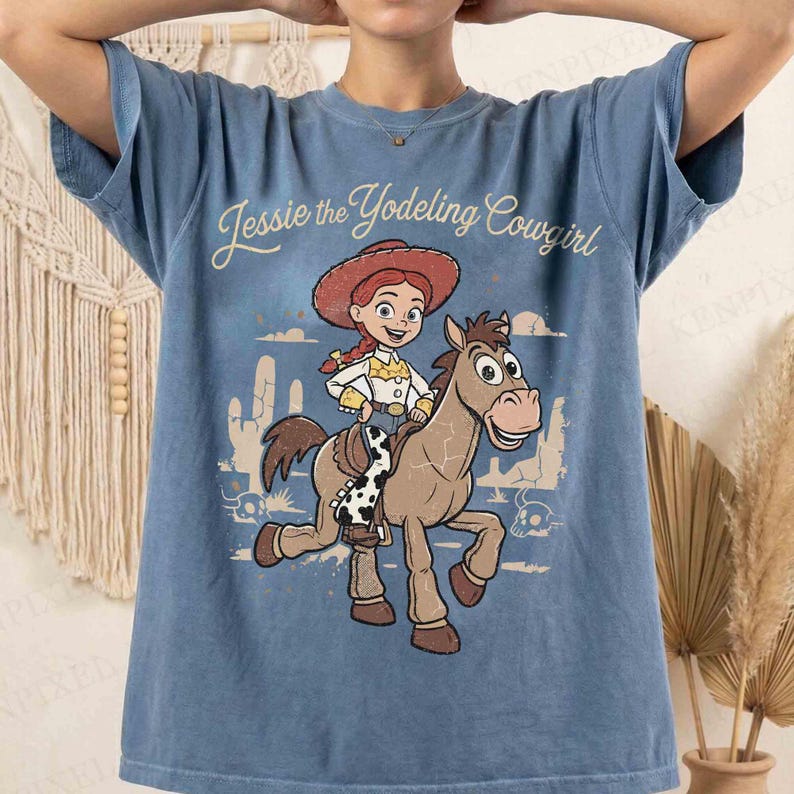 May include: A blue short-sleeve t-shirt featuring a graphic print of Jessie the Yodeling Cowgirl riding Bullseye the horse. The design includes a desert landscape with cacti and the text Jessie the Yodeling Cowgirl in a script font.