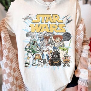 Puede incluir: Camiseta gr&aacute;fica de Star Wars de color crema con el logotipo de Star Wars y varios personajes de la franquicia. El dise&ntilde;o incluye personajes como Darth Vader, Yoda y Woody.