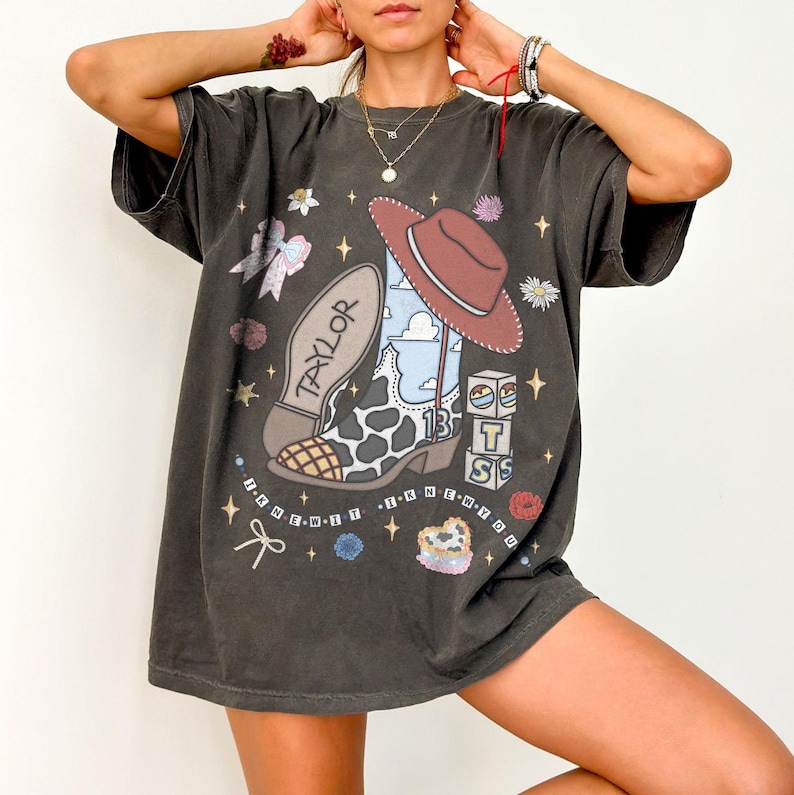 Puede incluir: Una camiseta gris carb&oacute;n de corte oversize con un estampado gr&aacute;fico de una bota vaquera con patr&oacute;n de manchas de vaca, un sombrero marr&oacute;n y bloques con las letras T, S y el n&uacute;mero 13. El texto TAYLOR e I KNEW IT I KNEW YOU est&aacute; impreso.