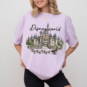 Puede incluir: Una camiseta lila con la palabra "Disneyworld" en cursiva. La camiseta presenta un gr&aacute;fico de un castillo, patos y un patito. La camiseta est&aacute; hecha de un material suave.