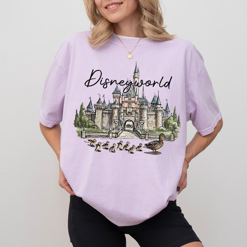 Puede incluir: Una camiseta lila con la palabra "Disneyworld" en cursiva. La camiseta presenta un gr&aacute;fico de un castillo, patos y un patito. La camiseta est&aacute; hecha de un material suave.