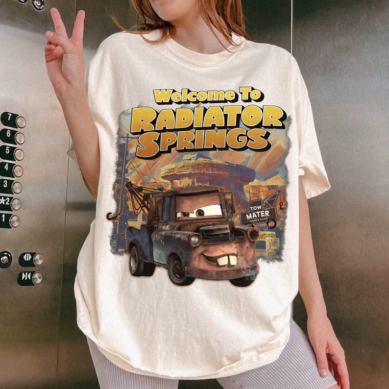 Pu&ograve; includere: T-shirt color crema con grafica di Cricchetto del film Cars, con la scritta "Welcome to Radiator Springs" in giallo e arancione. Il design include un'illustrazione dettagliata del personaggio e della citt&agrave;.