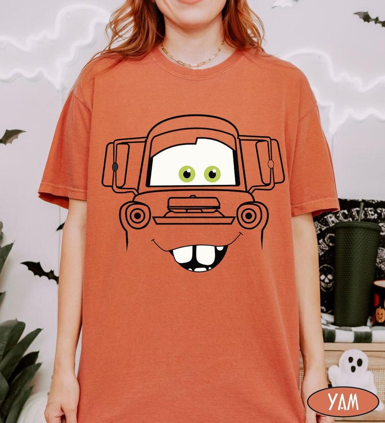 Op de afbeelding: Een roestoranje t-shirt met korte mouwen en een grote zwarte lijntekening van een cartoon-takelwagengezicht. De afbeelding bevat een voorruit met twee groene ogen, een grille en een brede mond met twee opvallende voortanden.