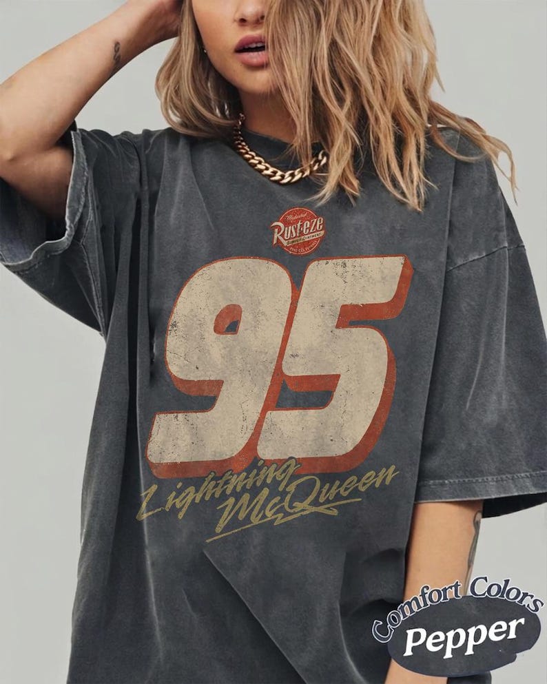 Puede incluir: Camiseta gris oscuro con un gr&aacute;fico desgastado que presenta el n&uacute;mero 95 en crema y rojo, las palabras "Lightning McQueen" y el texto "Rusteze". La camiseta est&aacute; etiquetada como "Comfort Colors Pepper".