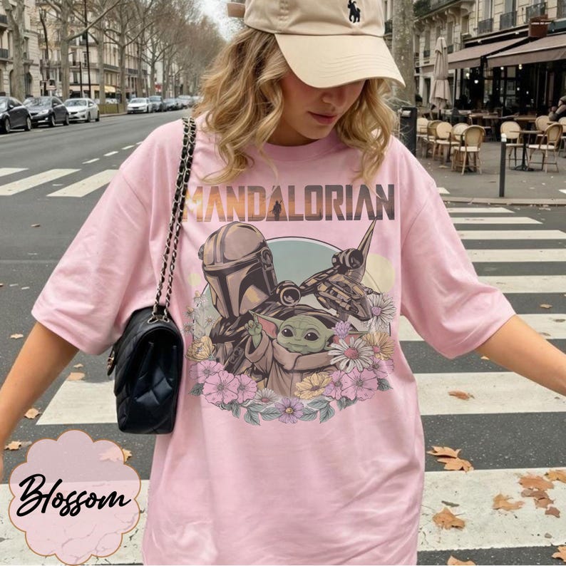 Puede incluir: Camiseta rosa claro con un dise&ntilde;o de The Mandalorian y Grogu, rodeados de flores, y la palabra "MANDALORIAN" en la parte superior. La camiseta es de corte holgado.