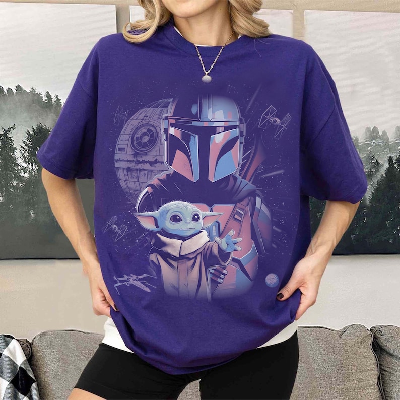 May include: A dark purple short-sleeve t-shirt featuring a graphic print of a helmeted character in armor standing behind a small green creature with large ears. The background includes a circular space station and small starships in space.
