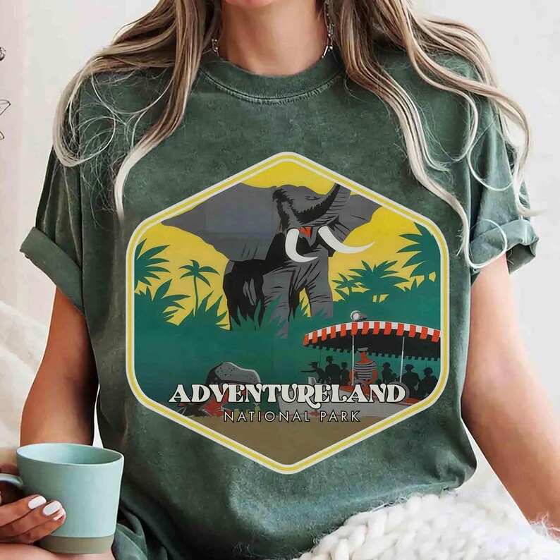 Op de afbeelding: Een donkergroen t-shirt met een zeshoekige grafische print van een olifant in een jungle en een boot met passagiers onder een rood-wit gestreepte luifel. De tekst ADVENTURELAND NATIONAL PARK staat in het wit onder de boot gedrukt.