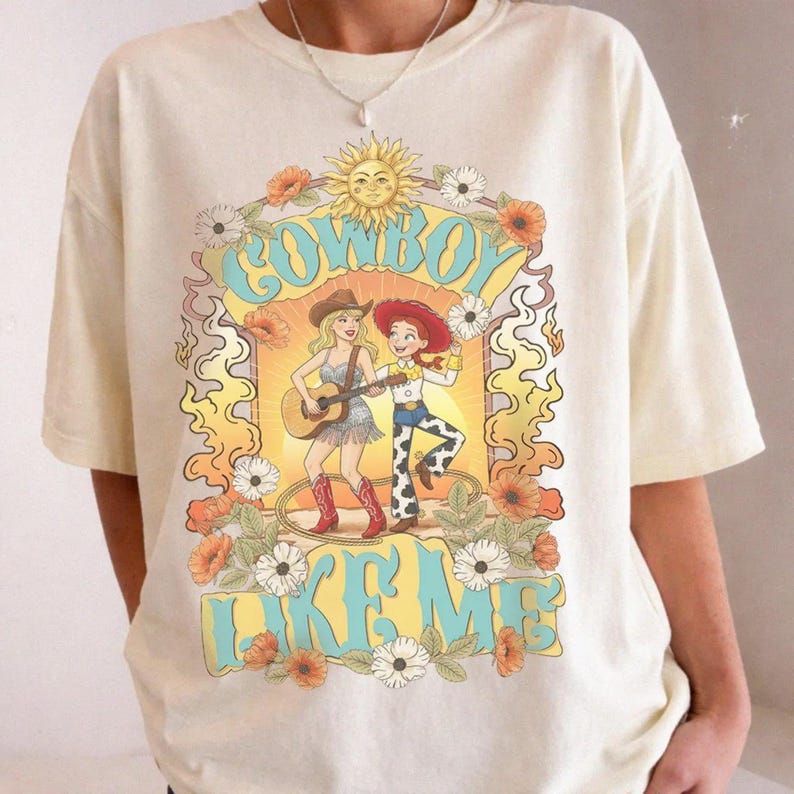 May include: A cream-colored short-sleeve t-shirt featuring a graphic print of two figures in cowboy attire, one playing a guitar. The design includes a sun, flowers, and the text "COWBOY LIKE ME" in a light blue font, surrounded by orange flames.