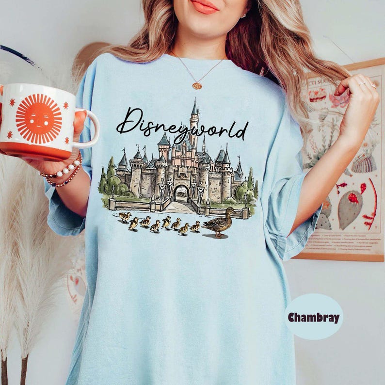 Puede incluir: Camiseta azul claro con un gr&aacute;fico del castillo de Disneyworld y el texto "Disneyworld". El gr&aacute;fico incluye un castillo, patos y la palabra "Disneyworld". Una taza roja y blanca con un dise&ntilde;o de sol se sostiene en la mano.