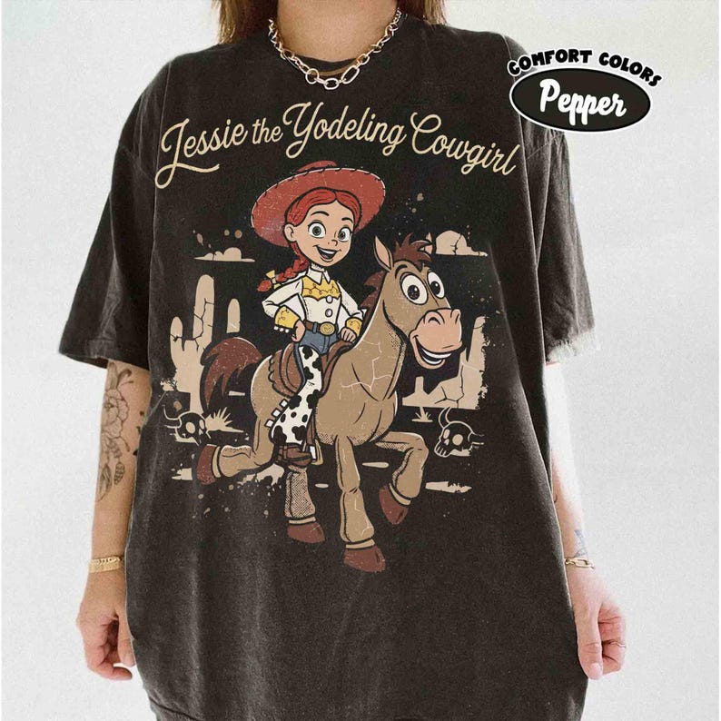 May include: A charcoal gray short-sleeve t-shirt featuring a graphic of a cowgirl riding a horse. The text reads Jessie the Yodeling Cowgirl. The design includes a desert landscape with cacti and skulls. A label in the corner says Comfort Colors Pepper.