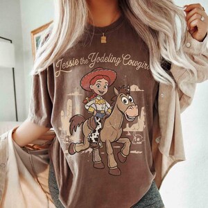 May include: A brown short-sleeve t-shirt featuring a graphic print of Jessie the Yodeling Cowgirl riding Bullseye the horse. The design includes a desert landscape with cacti and the text Jessie the Yodeling Cowgirl in a cursive font.