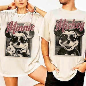 May include: Off-white t-shirts featuring a black and white image of Mickey and Minnie Mouse wearing sunglasses. The word "Minnie" is printed above Minnie's image in a maroon script, and "Mickey" is printed above Mickey's image.