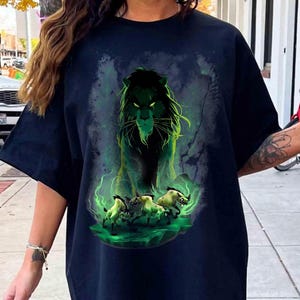 Puede incluir: Camiseta negra con una ilustraci&oacute;n verde y negra de un le&oacute;n con ojos y melena brillantes, rodeado de niebla verde. Debajo del le&oacute;n, tres &ntilde;us corriendo, tambi&eacute;n verdes. El dise&ntilde;o se presenta sobre un fondo oscuro.