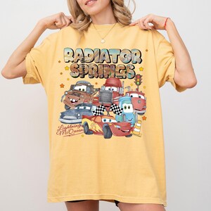 May include: Yellow t-shirt featuring a graphic of characters from the movie Cars, including Lightning McQueen, Mater, and others, with the text "Radiator Springs" and "Lightning McQueen". The design includes stars and a traffic light.