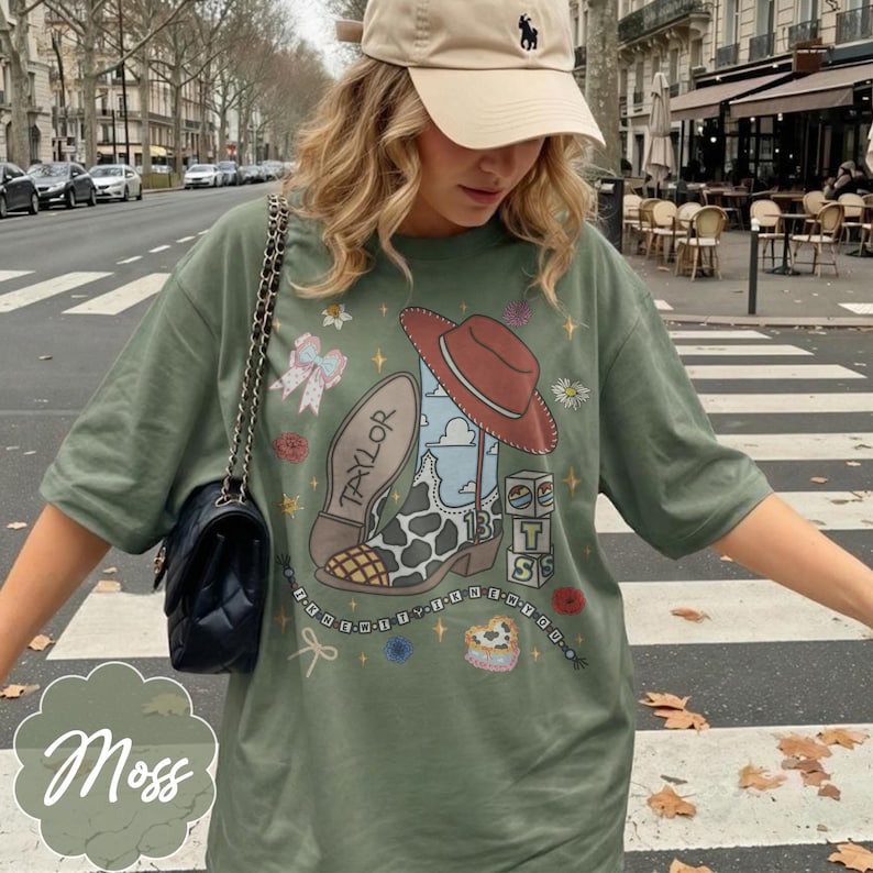 Puede incluir: Una camiseta verde oliva con un estampado gr&aacute;fico de una bota de vaquero con manchas de vaca, un sombrero rojo, el nombre Taylor, el n&uacute;mero 13 y bloques con las letras T, S y un coraz&oacute;n. El dise&ntilde;o incluye una cadena de cuentas y flores.