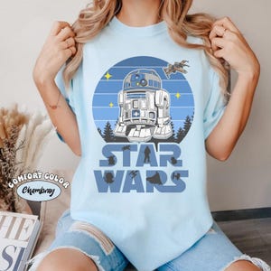 Op de afbeelding: Lichtblauw T-shirt met een Star Wars-ontwerp. De afbeelding bevat R2-D2, een ruimteschip en de woorden "STAR WARS" in blauw. Het shirt heeft ook de woorden "COMFORT COLOR Chambray" aan de linkerkant.