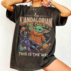 May include: Dark gray t-shirt featuring a graphic from The Mandalorian. The design includes the text "The Mandalorian The Child" with an illustration of Grogu and the Mandalorian. The phrase "This is the Way" is also displayed.