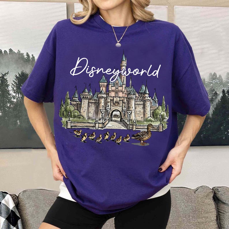 Puede incluir: Una camiseta morada con un gr&aacute;fico de Disney World. El gr&aacute;fico incluye un castillo, patos y las palabras "Disneyworld" en una fuente de escritura. La camiseta es de color liso y tiene mangas cortas.