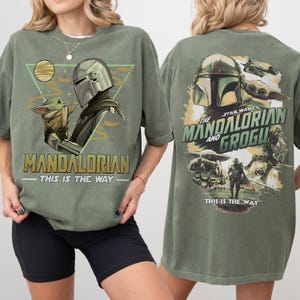 K&ouml;nnte beinhalten: Olivgr&uuml;nes T-Shirt mit einer Grafik von The Mandalorian und Grogu. Das Frontdesign enth&auml;lt den Text "MANDALORIAN" und "THIS IS THE WAY". Die R&uuml;ckseite des Shirts zeigt "THE MANDALORIAN AND GROGU" mit zus&auml;tzlichen Bildern.