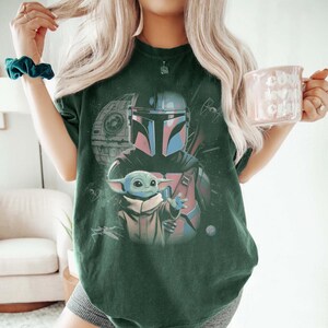 May include: A dark green short-sleeve t-shirt featuring a graphic print of a Mandalorian character in a helmet and a small green creature with large ears. The design includes a space station and starships in the background with a vintage aesthetic.