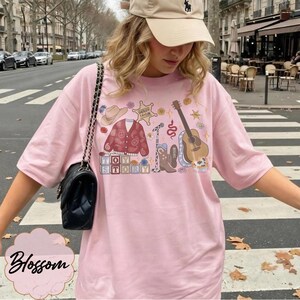 May include: A light pink t-shirt featuring a graphic print of a cowboy hat, a paisley cardigan, a sheriff badge labeled Sheriff Taylor, a microphone, a snake, a guitar, a cowboy boot, and blocks spelling Toy Story with floral accents.
