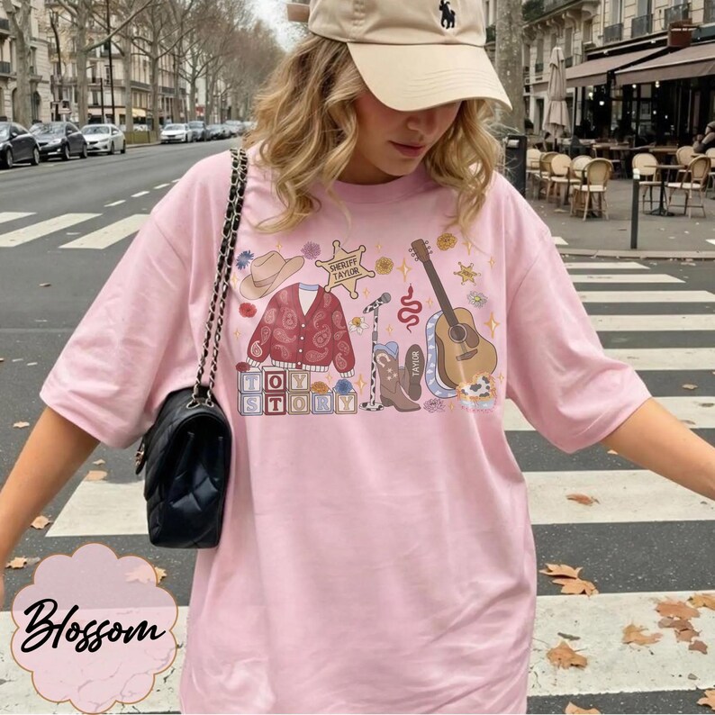 May include: A light pink t-shirt featuring a graphic print of a cowboy hat, a paisley cardigan, a sheriff badge labeled Sheriff Taylor, a microphone, a snake, a guitar, a cowboy boot, and blocks spelling Toy Story with floral accents.