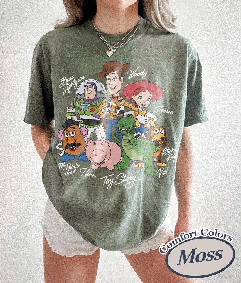 May include: An olive green t-shirt with a graphic of Toy Story characters, including Woody and Buzz Lightyear. The shirt features the text "Toy Story" and "Comfort Colors Moss".