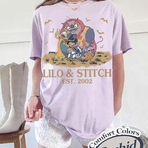 May include: A light purple t-shirt featuring a graphic of characters from Lilo & Stitch, with the text "Lilo & Stitch EST. 2002". The shirt is a Comfort Colors brand in the color Orchid.