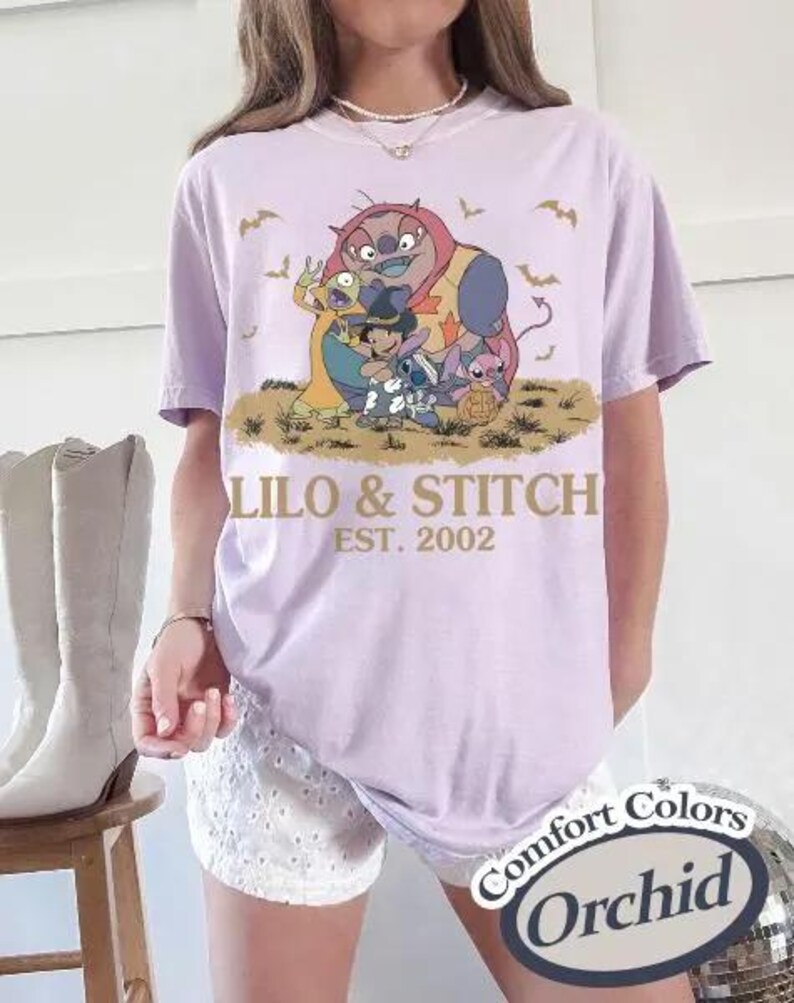 May include: A light purple t-shirt featuring a graphic of characters from Lilo & Stitch, with the text "Lilo & Stitch EST. 2002". The shirt is a Comfort Colors brand in the color Orchid.