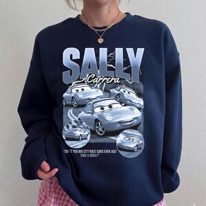 Puede incluir: Sudadera azul marino con un dise&ntilde;o de Sally Carrera de la pel&iacute;cula Cars. El dise&ntilde;o incluye el texto "SALLY Carrera" y la cita "DON'T YOU BIG CITY RACE CARS EVER JUST TAKE A DRIVE?"
